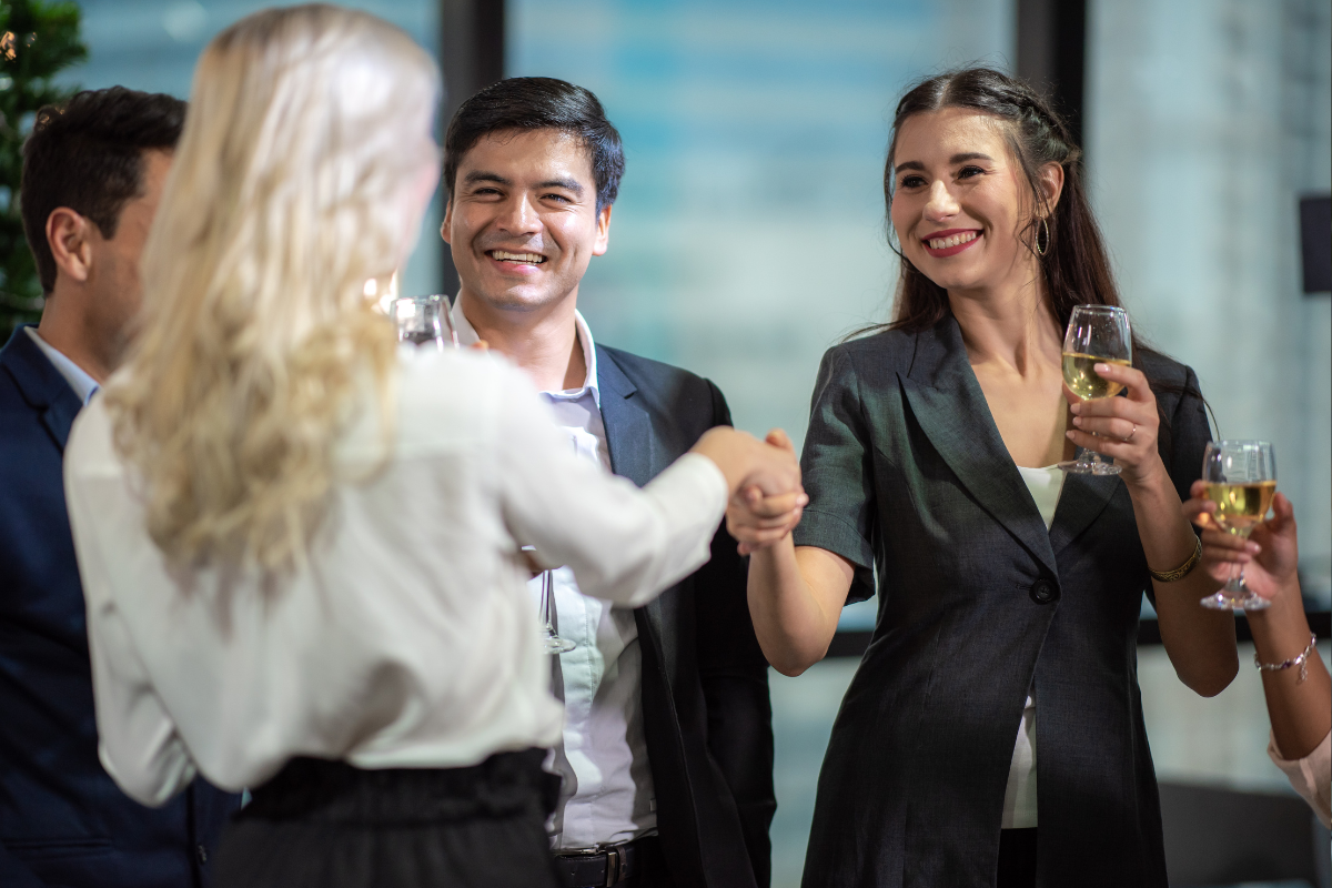 A group of professionals networking at an office event, smiling and shaking hands while holding glasses of wine.