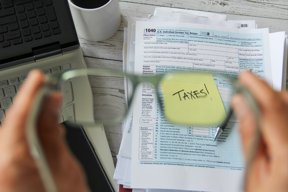 Person holding eyeglasses while focusing on tax forms with a sticky note that says ‘TAXES!’ on a desk with a laptop and coffee cup.